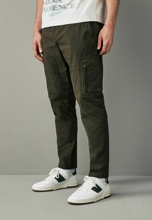 Olivgrüne Cargo-Hosen mit Seitentaschen, strukturierter Stoff, schmal zulaufender Schnitt, getragen mit weißen Sneakers mit grünen Akzenten.