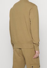 Brun sweatshirt med ribbade muddar och fåll, långa ärmar och rund halsringning, tillverkad av mjukt bomullsmaterial. Bärs med matchande byxor.