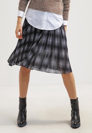 Kvinde iført sort-hvid ternet plisseret nederdel, brun sweater, hvid skjorte og sorte ankelstøvler stående med benene fra hinanden.