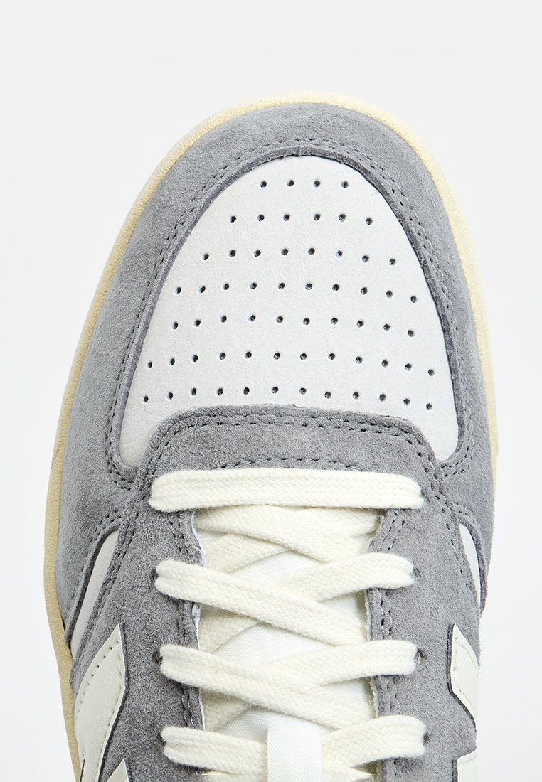 Zapatilla de ante gris y piel blanca con caja de dedos perforada, cordones cremosos y suela de color claro; presenta un destacado detalle de marca en el lado.