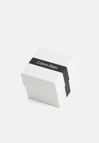 Boîte carrée blanche avec une bande noire comportant le texte "Calvin Klein". La boîte a une finition lisse et des bords nets, avec un design minimaliste.