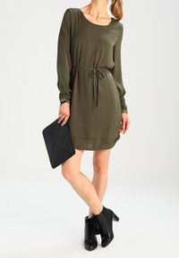 Vestido largo de manga larga en verde oliva con cintura anudada. Combinado con botas negras de tobillo y un bolso de mano geométrico negro. Textura suave.