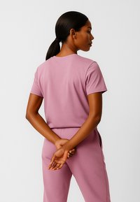 Kobieta o ciemnych włosach związanych nisko w kucyk, ubrana w różowy zestaw składający się z krótkiego rękawa i spodni, stojąca z rękami splecionymi za plecami.