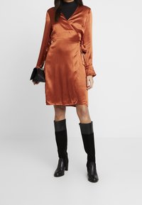 Satin-kääntömekko palaneessa oranssissa, pitkät hihat röyhelöillä, yhdistettynä polven yli ulottuviin mustiin saappaisiin, joissa on nahka- ja samettipinta.