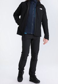 Svart vattentät jacka med huva, har en dragkedja framtill och blå accenter; kombinerad med matchande svarta byxor och stövlar.