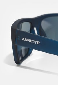 Modré plastové slnečné okuliare s tmavými sklami, s obdĺžnikovým tvarom a strieborným logom "ARNETTE" na strane.