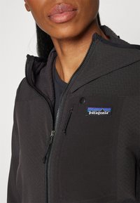 Giacca in pile nera foderata con zip frontale, colletto alto e tasche laterali. Presenta un logo Patagonia sul lato sinistro del petto.