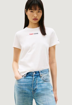 Joven mujer vistiendo una camiseta blanca "Tommy Jeans" y jeans azules, posando con una mano en el bolsillo contra un fondo liso.