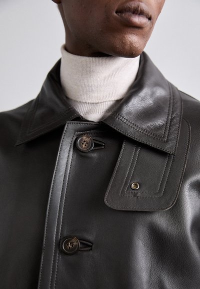 Mørk læderjakke med bred krave, der har knaplukning og dekorativ syning. Båret over en lysfarvet mock turtleneck.