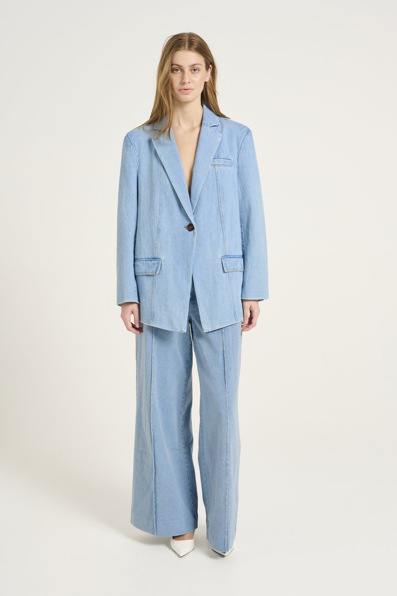 Blazer oversized en denim bleu clair et pantalon à jambe large, présentant des détails de couture minimalistes et une fermeture à bouton marron unique.