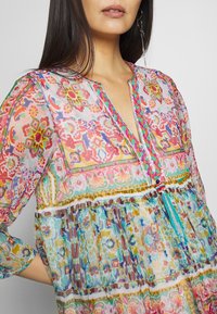 Blusa colorida con estampados, mangas largas, escote en V y ribete trenzado; presenta diseños florales y geométricos en rosa, azul y verde.