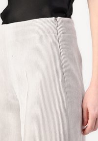 Lys grå corduroy bukser med ribbet tekstur, detaljer med lynlås i siden og lige ben design. Nærbillede af stoffet og pasformen.