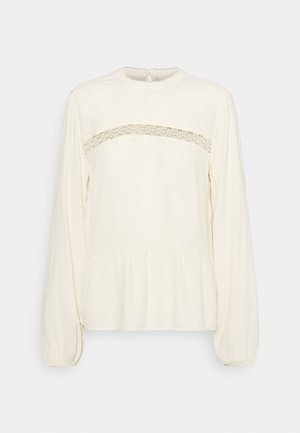 Cream-colored long-sleeve blouse with a round neckline, featuring a lace panel across the chest and a gathered peplum hem. Soft texture.