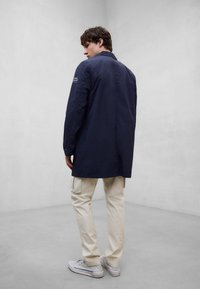 Ein Mann, der mit dem Rücken zum Betrachter steht, trägt einen marineblauen langen Mantel, beige Cargohosen und weiße Turnschuhe und steht in einem schlichten grauen Raum.