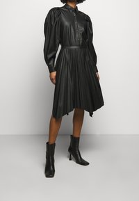 Robe noire plissée en simili cuir, avec des manches longues bouffantes, un devant à boutons et un ourlet asymétrique jusqu'aux genoux.