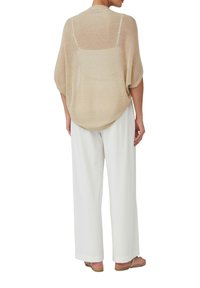 Beige gebreide oversized top met een losse pasvorm, voorzien van een doorzichtig achterpand, in combinatie met witte wijdlopende broek en tan sandalen.