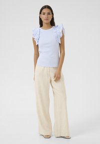 Haut bleu clair à côtes avec des accents de manches volantes, associé à un pantalon ample beige. La tenue présente une coupe décontractée et des textures douces.