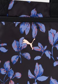 Μαύρο ύφασμα με μεγάλο μπλε και ροζ λουλουδάτο σχέδιο και μικρή λεπτομέρεια με ροζ χρυσό λογότυπο Puma κοντά σε φερμουάρ.