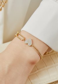 Guldtonad kedjearmband med ett hjärtformigt hänge, prytt med klara rhinstenar. Har en låsning och en texturerad länkdesign.