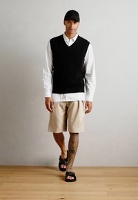 Svart strikket vest over en hvit skjorte med krage, beige shorts og svarte sandaler. Har en enkel svart caps og synlige tatoveringer på leggen.