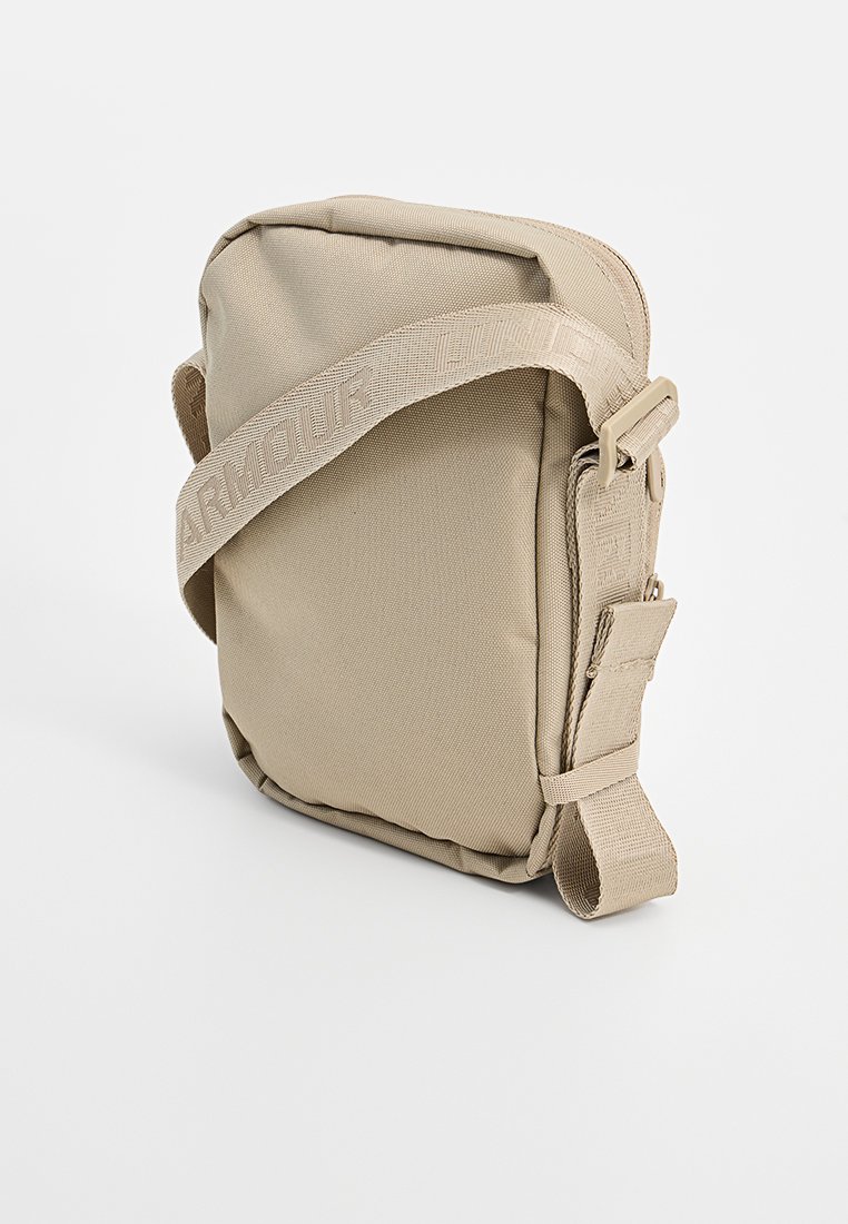Sac à bandoulière en tissu beige avec fermeture éclair, bretelle réglable et logo en relief sur la bretelle ; forme rectangulaire avec texture lisse.