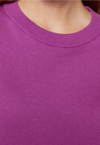 Närbild av en person som bär en lila stickad tröja med rund halsringning och ribbat mönster, som visar halsen och övre bröstområdet.