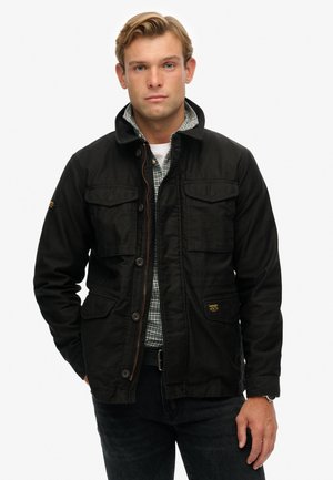 Man in een zwarte knopenjacket over een groen geruit shirt, staand tegen een eenvoudige witte achtergrond, met zijn handen in zijn zakken.