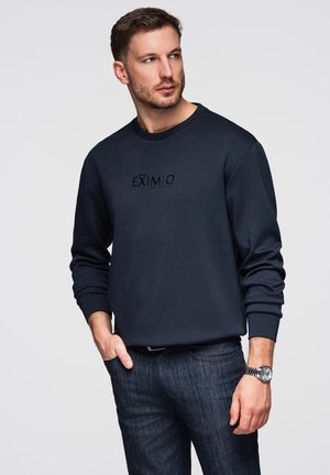 Mies, jolla on päällään laivastonsininen collegepaita, jossa teksti "EXIMIO", tummat farkut ja rannekello, seisoo käsi taskussa ja katsoo sivulle.