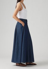 Džínová maxi sukně v tmavě modré barvě se bočními kapsami, vysokým pasem a viditelným švem. Nositá s bílým tílkem a plochými sandály.