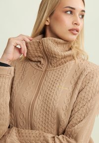 Une jeune femme ajuste le col haut d'un pull zippé texturé beige, regardant sur le côté contre un fond clair.