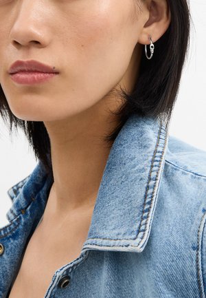 Silver hoop earring featuring a sleek design with a shiny surface. Worn with a light blue denim jacket with a classic collar and visible stitching.