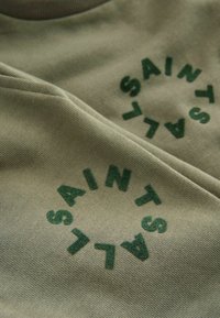 Olivenfarbener Baumwoll-Hoodie mit rundem "SAINTS"-Logo in dunkelgrün, strukturiertem Stoff und einer weichen, glatten Oberfläche.