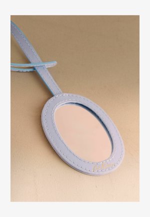 Ovalno ročno ogledalo z rahlo modrim všitim okvirjem in trakom, ki odseva bež površino, postavljeno na ujemajoči se bež ozadje.