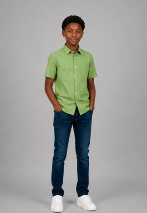Adolescent debout, les mains dans les poches, portant une chemise verte à manches courtes, un jean bleu foncé et des baskets blanches sur un fond gris uni.