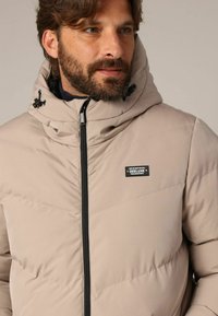 Beige gewatteerd jack met een hoge kraag, zwarte ritsluiting en een logopatch op de borst. Heeft een doorgestikte textuur en een verstelbare capuchon.