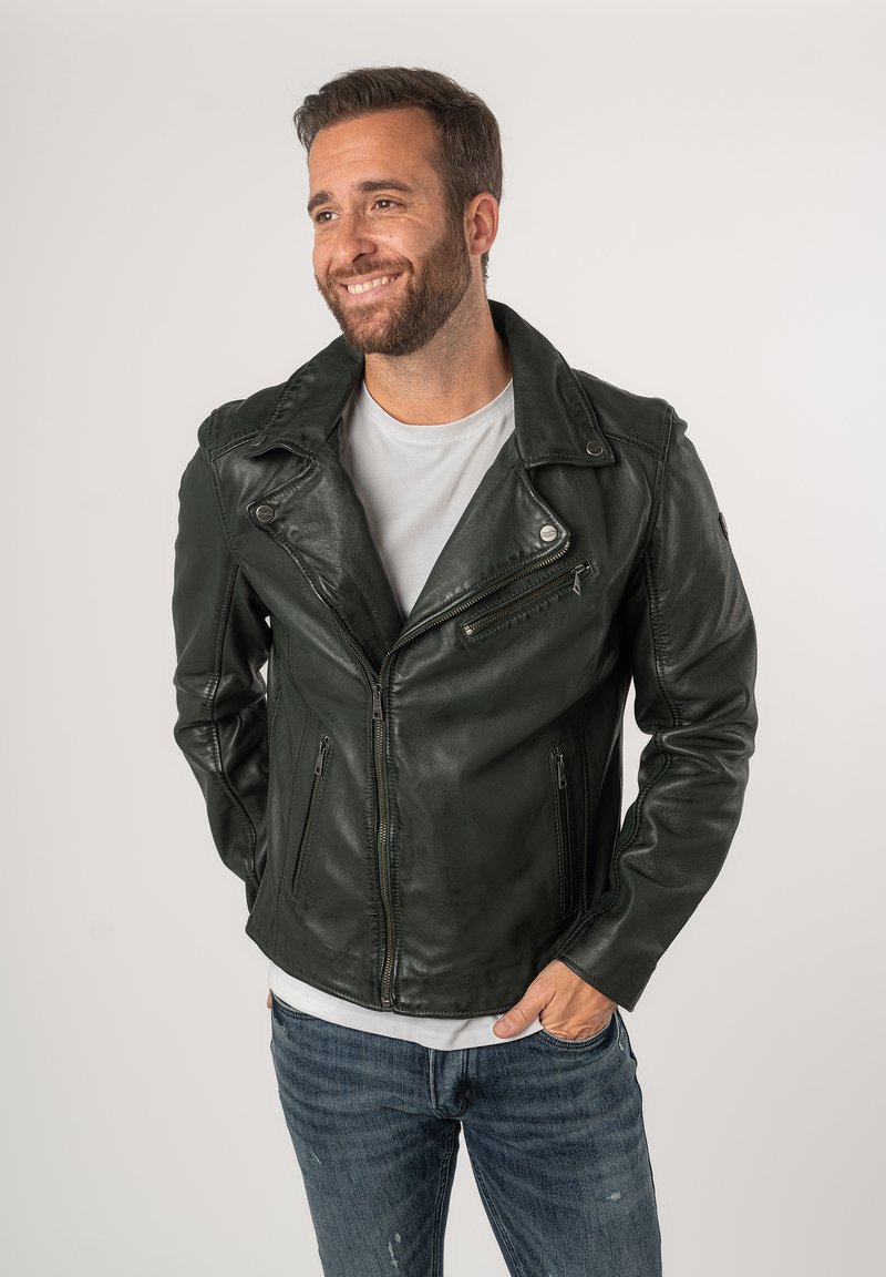 Giacca da biker in pelle verde scuro con colletto a punte, tasche con zip e design aderente. Indossata sopra una maglietta grigia chiara.