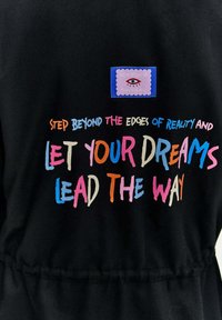 Melnas auduma ar krāsainu tekstu "Pārlieciet pāri realitātes malām un ļaujiet sapņiem vadīt ceļu," kā arī uzšuvums ar acs grafiku.