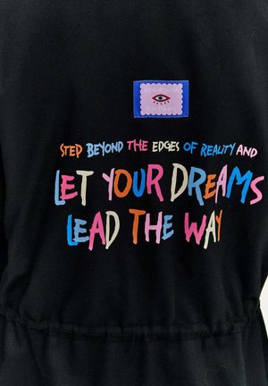 Černá tkanina s barevným textem, který zní "Překročte hranice reality a nechte své sny vést vás," plus nášivka s grafikou oka.
