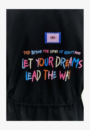 Černá tkanina s barevným textem, který zní "Překročte hranice reality a nechte své sny vést vás," plus nášivka s grafikou oka.