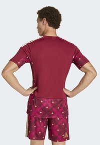 Maroon atletski dres i šorts s geometrijskim uzorcima u raznim nijansama ružičaste, sa zlatnim akcentima na bočnoj strani šortsa.