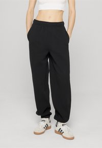 Asmuo dėvi juodus elastiniu juosmeniu sportines kelnes, rankos kišenėse, baltą trumpą marškinėlį ir baltus Adidas sportbačius su juodomis juostelėmis.