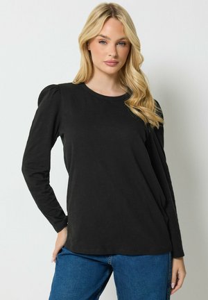 Mujer rubia con ondas sueltas que lleva una blusa negra de manga larga con hombros abullonados y vaqueros azules, de pie sobre un fondo blanco.