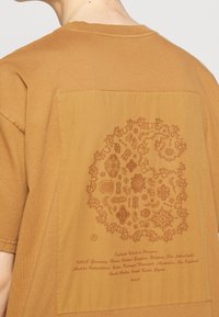 Person wearing a brown t-shirt with an intricate paisley pattern shaped like a stylized "C" and printed text listing various countries.