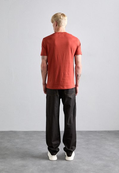 Red short-sleeve t-shirt, fitted design. Paired with loose-fitting black pants, featuring orange stitching. Plain white sneakers.