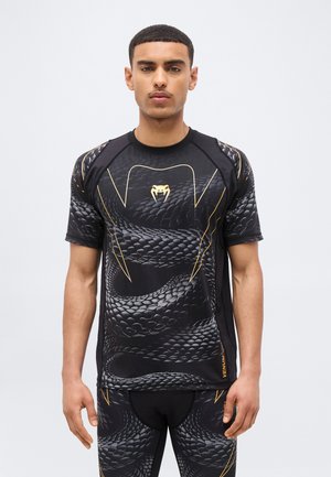 Man wearing black athletic shirt and leggings with snake scale pattern and gold snake logo on chest against plain background.