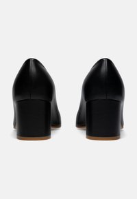 Chaussures à talons hauts en cuir noir avec un bout pointu et un large talon bloc. Texture lisse et design simple sans embellissements visibles.