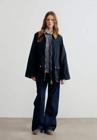 Νεαρό άτομο που φοράει μαύρο, φαρδύ μπουφάν με καφέ γιακά, ριγέ πουκάμισο, φαρδιά σκούρα παντελόνια και γκρι παπούτσια, στέκεται μπροστά σε απλό φόντο.