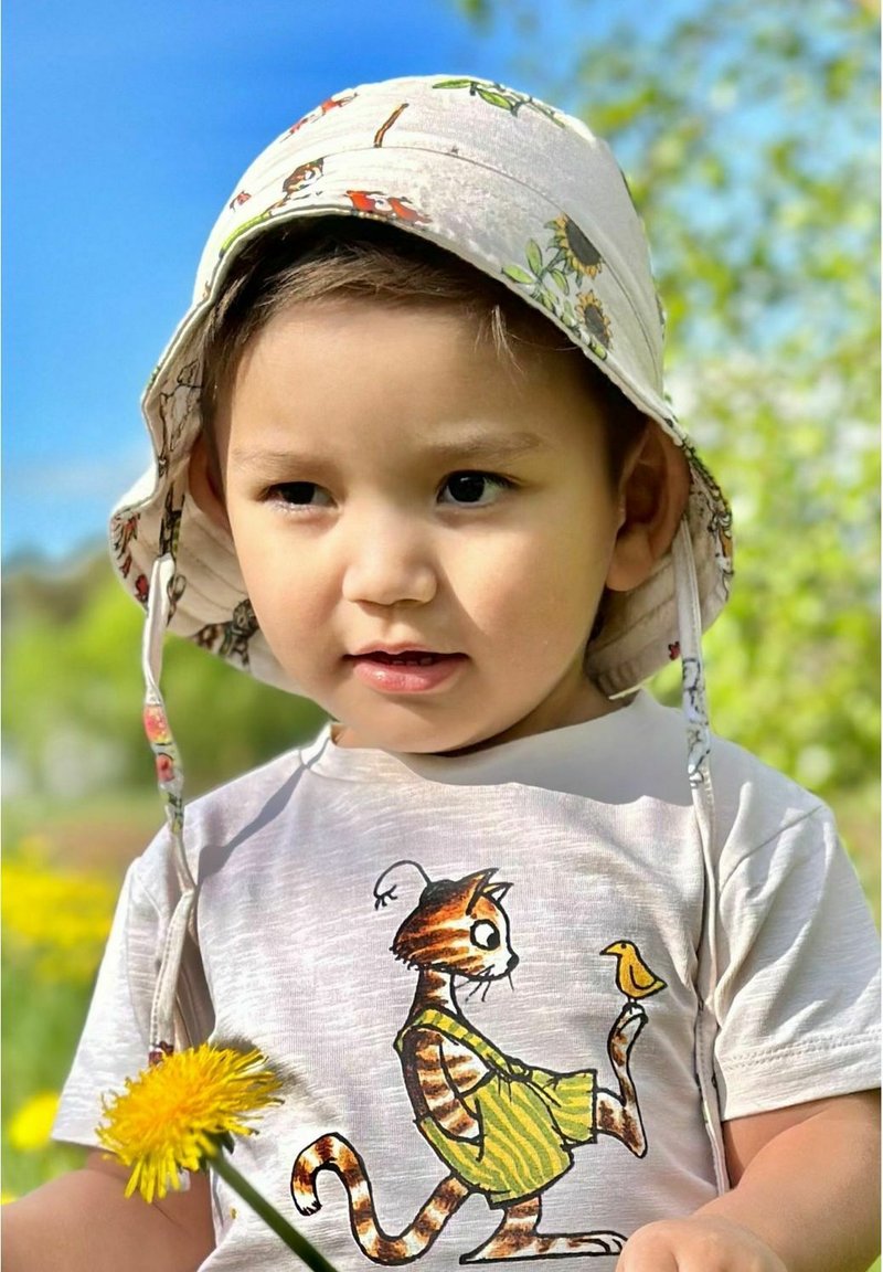 Un enfant portant un chapeau à larges bords à motifs et un t-shirt clair représentant un chat de dessin animé tenant un oiseau, sur un fond vert.