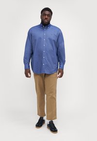 Blue button-up shirt with long sleeves, cotton material, chest logo detail, paired with tan pants and black sneakers.