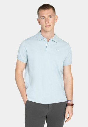 Joven vestido con una camiseta tipo polo de manga corta azul claro y pantalones oscuros, de pie con una mano en el bolsillo frente a un fondo liso.